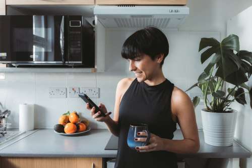 Woman Stands In The Kitchen And Smiles While Using Cell Phone Photo
